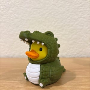 Puede incluir: Un pato de goma amarillo con pico naranja que lleva un disfraz de cocodrilo verde oscuro. El disfraz presenta un exterior texturizado con escamas, una barriga blanca y una capucha con forma de cabeza de cocodrilo abierta con dientes blancos.