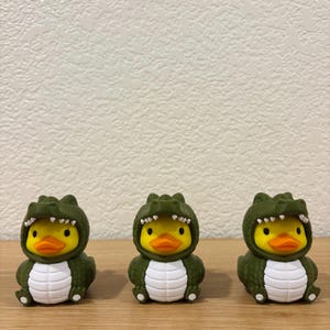 Puede incluir: Tres patos de goma amarillos con disfraces de caim&aacute;n verde oscuro. Cada pato tiene un pico naranja brillante, ojos negros y una barriga blanca. Las capuchas de caim&aacute;n muestran dientes blancos y una piel verde texturizada sobre la cabeza.