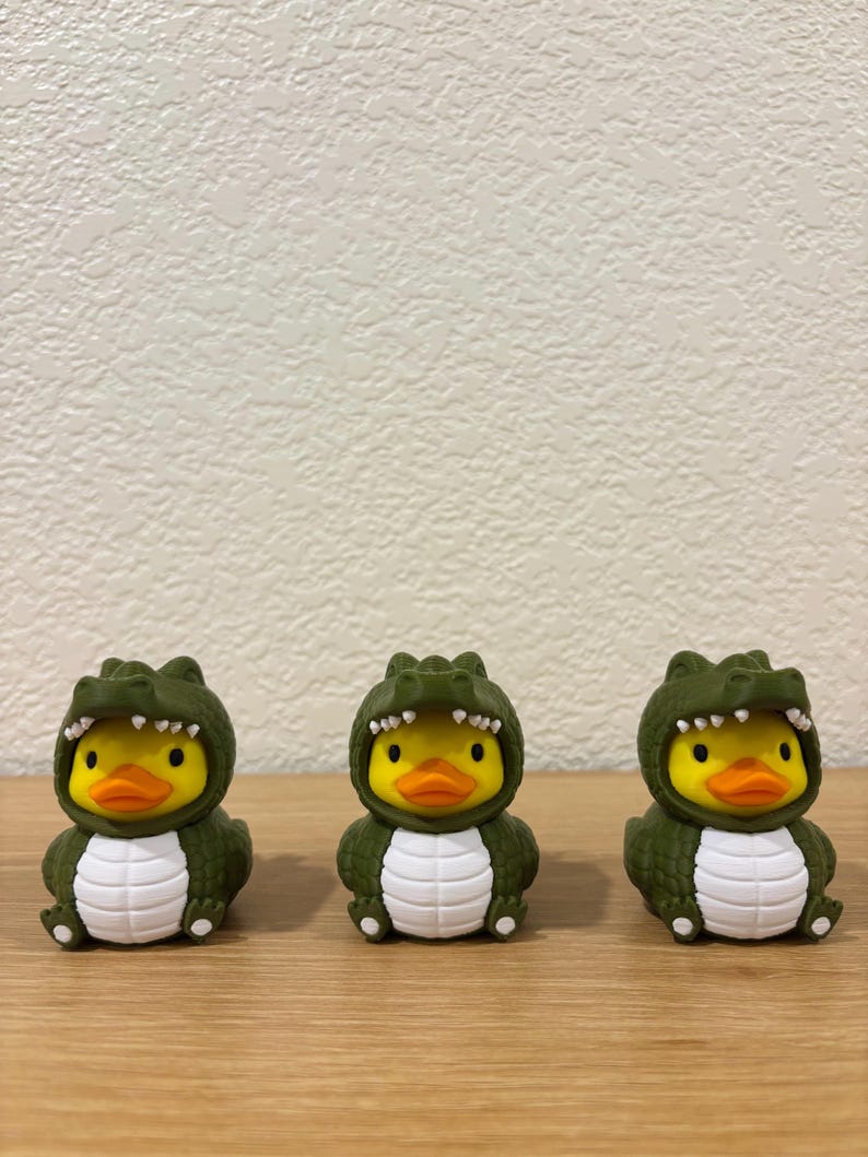 Puede incluir: Tres patos de goma amarillos con disfraces de caim&aacute;n verde oscuro. Cada pato tiene un pico naranja brillante, ojos negros y una barriga blanca. Las capuchas de caim&aacute;n muestran dientes blancos y una piel verde texturizada sobre la cabeza.