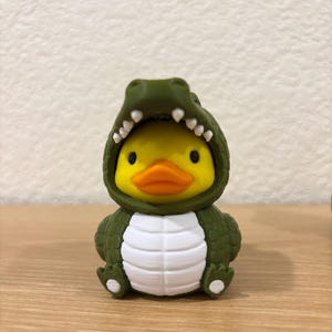 Puede incluir: Un pato de goma amarillo con un disfraz de caim&aacute;n verde oliva oscuro. El disfraz presenta un exterior texturizado con escamas, un panel abdominal blanco y una capucha abierta que revela la cara del pato con peque&ntilde;os dientes blancos.
