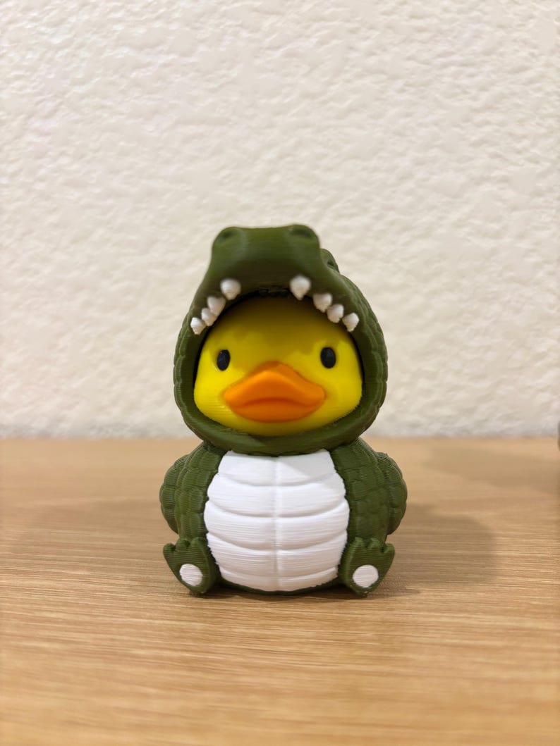Puede incluir: Un pato de goma amarillo con un disfraz de caim&aacute;n verde oliva oscuro. El disfraz presenta un exterior texturizado con escamas, un panel abdominal blanco y una capucha abierta que revela la cara del pato con peque&ntilde;os dientes blancos.