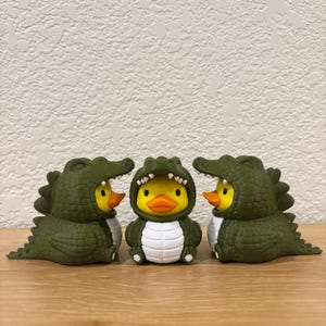 Puede incluir: Tres patos de goma amarillos vestidos con disfraces de caim&aacute;n de color verde oliva oscuro. Los disfraces presentan escamas texturizadas, dientes blancos a lo largo de la abertura de la capucha y un panel blanco en el vientre. Los patos tienen picos naranjas y ojos negros.