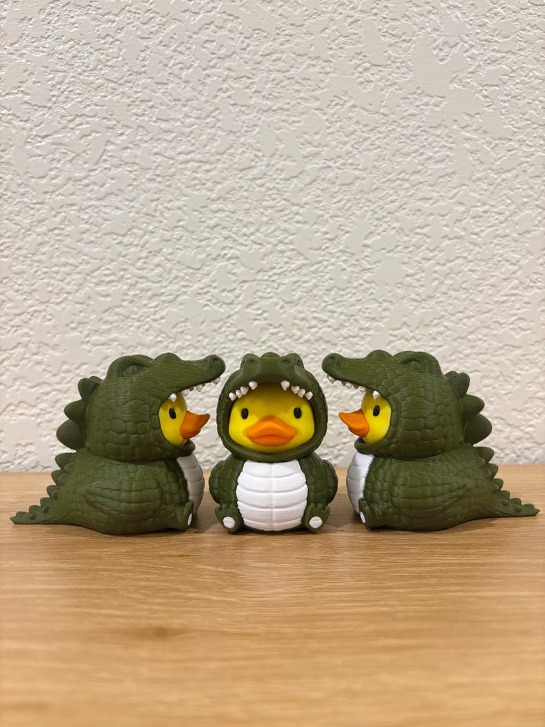 Puede incluir: Tres patos de goma amarillos vestidos con disfraces de caim&aacute;n de color verde oliva oscuro. Los disfraces presentan escamas texturizadas, dientes blancos a lo largo de la abertura de la capucha y un panel blanco en el vientre. Los patos tienen picos naranjas y ojos negros.