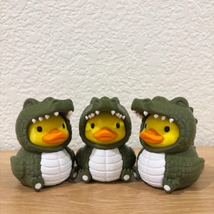 Puede incluir: Tres patos de goma amarillos con disfraces de caim&aacute;n de color verde oliva oscuro. Cada disfraz presenta un exterior texturizado con escamas, un panel blanco en el vientre y una capucha con dientes blancos que enmarcan la cara del pato.