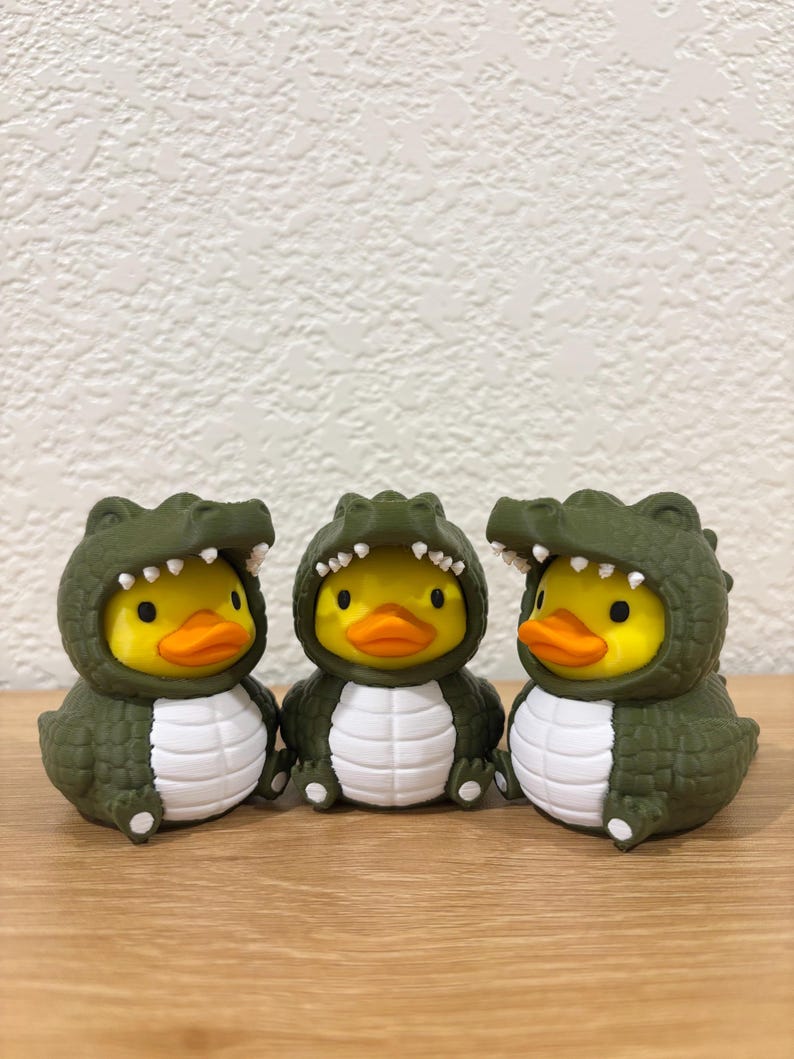 Puede incluir: Tres patos de goma amarillos con disfraces de caim&aacute;n de color verde oliva oscuro. Cada disfraz presenta un exterior texturizado con escamas, un panel blanco en el vientre y una capucha con dientes blancos que enmarcan la cara del pato.