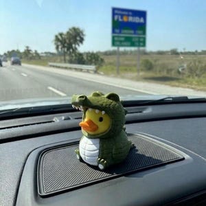 Puede incluir: Una peque&ntilde;a figura de pato de goma amarillo con un disfraz de caim&aacute;n verde, vientre blanco y escamas texturizadas. El pato tiene un pico naranja y est&aacute; colocado sobre una superficie negra texturizada del salpicadero dentro de un veh&iacute;culo.