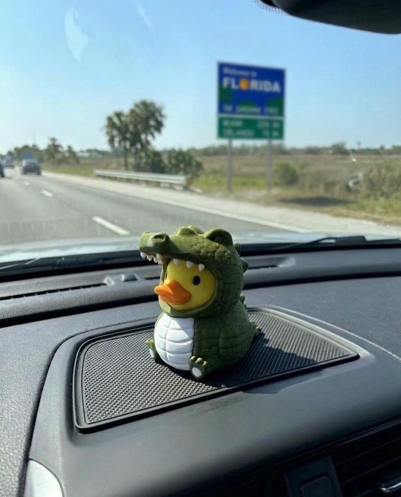 Puede incluir: Una peque&ntilde;a figura de pato de goma amarillo con un disfraz de caim&aacute;n verde, vientre blanco y escamas texturizadas. El pato tiene un pico naranja y est&aacute; colocado sobre una superficie negra texturizada del salpicadero dentro de un veh&iacute;culo.