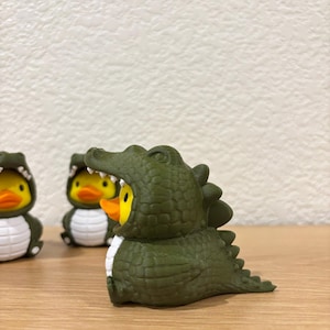 Puede incluir: Tres peque&ntilde;os patos de goma, cada uno con un disfraz de caim&aacute;n de color verde oliva oscuro. El disfraz presenta un exterior texturizado y escamoso, una hilera de dientes blancos alrededor de la cara y una cola con p&uacute;as.
