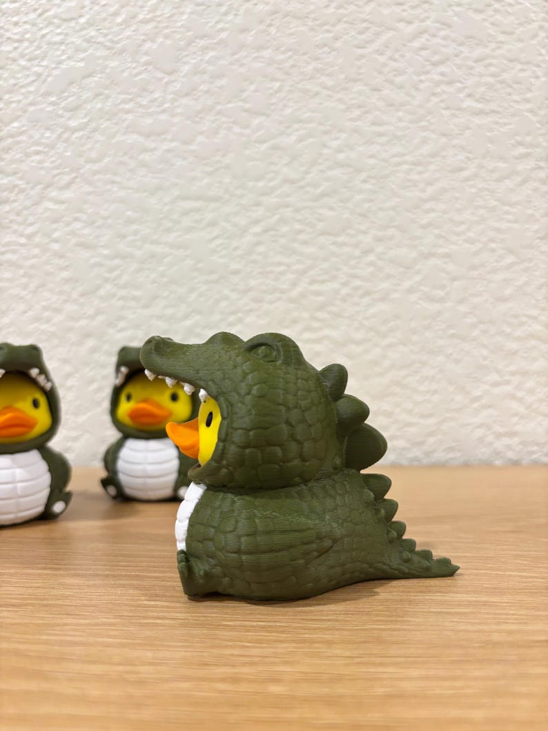 Puede incluir: Tres peque&ntilde;os patos de goma, cada uno con un disfraz de caim&aacute;n de color verde oliva oscuro. El disfraz presenta un exterior texturizado y escamoso, una hilera de dientes blancos alrededor de la cara y una cola con p&uacute;as.