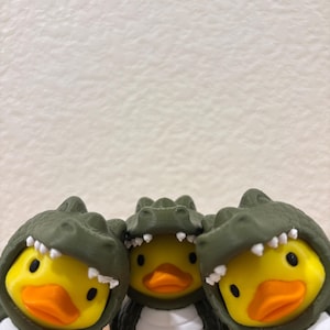 Puede incluir: Tres patos de goma amarillos con disfraces de caim&aacute;n de color verde oliva oscuro. Cada disfraz presenta una capucha texturizada con escamas y dientes blancos puntiagudos que enmarcan la cara del pato, y un vientre blanco con crestas horizontales.