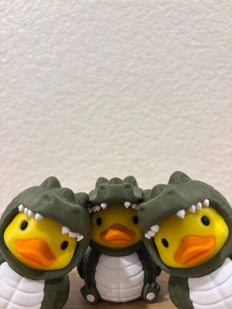 Puede incluir: Tres patos de goma amarillos con disfraces de caim&aacute;n de color verde oliva oscuro. Cada disfraz presenta una capucha texturizada con escamas y dientes blancos puntiagudos que enmarcan la cara del pato, y un vientre blanco con crestas horizontales.
