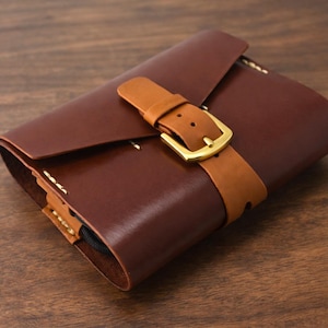May include: A handcrafted, dark brown leather wallet with a lighter brown leather strap and a gold-colored buckle. The wallet is closed with a flap and stitched with light-colored thread. The product is displayed on a wooden surface.