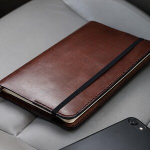 May include: A rectangular notebook with a smooth, dark brown leather cover and a black elastic band closure. The notebook features visible stitching along the edges and a small, light grey fabric ribbon bookmark extending from the side of the pages.