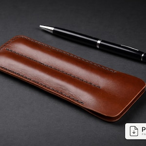 May include: A brown leather pen case with black stitching and a black pen with silver accents. The pen case is rectangular and has a smooth, glossy finish. The pen is lying on a dark gray surface.