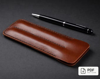 May include: A brown leather pen case with black stitching and a black pen with silver accents. The pen case is rectangular and has a smooth, glossy finish. The pen is lying on a dark gray surface.