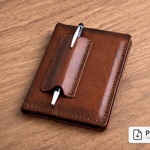 May include: A brown leather notebook with a pen holder and a silver pen inserted. The notebook is rectangular and has a stitched border. The background is a wooden surface.