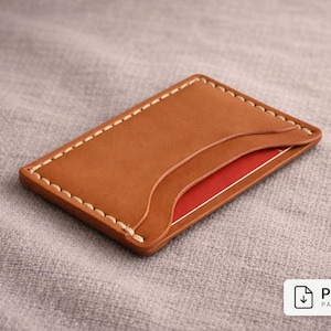 Puede incluir: Un tarjetero de cuero marrón hecho a mano con un diseño minimalista. La cartera tiene un bolsillo en la parte delantera y una ranura para tarjetas. Los bordes están cosidos con hilo blanco. Una tarjeta roja es parcialmente visible.