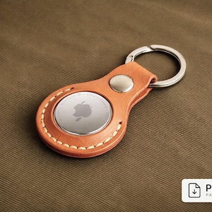 May include: A tan leather keychain holder with a silver Apple AirTag. The holder is stitched with white thread and has a silver metal keyring. The background is a brown fabric.