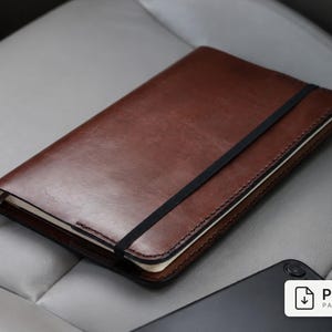 May include: A brown leather notebook cover featuring visible stitching along the edges and a black elastic band closure. A thin gray ribbon bookmark extends from the side. A white rectangular label with the text PDF PATTERN is visible.