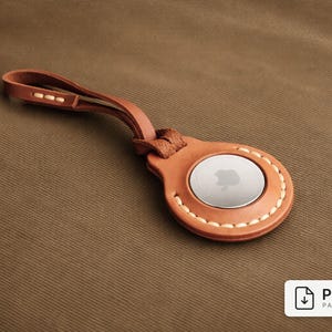 Puede incluir: Un soporte de cuero marrón para AirTag con un lazo, con un disco plateado con el logotipo de Apple. El soporte está cosido con hilo blanco y descansa sobre una superficie de tela marrón. La imagen incluye un icono de patrón PDF.
