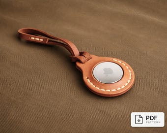May include: A brown leather AirTag holder with a loop, featuring a silver disc with the Apple logo. The holder is stitched with white thread and rests on a brown fabric surface. The image includes a PDF pattern icon.