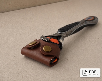 May include: A brown leather razor cover with two brass-colored snap buttons, fitted over the head of a black and silver multi-blade razor. The cover is designed to protect the blade cartridge. A small white rectangular label reads PDF PATTERN.