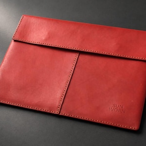 Può includere: Una custodia per documenti in pelle rossa con patta ripiegabile e cuciture visibili lungo i bordi. La parte anteriore presenta una cucitura verticale e il testo SUN WORN in rilievo nell'angolo in basso a destra. La superficie ha una finitura opaca liscia.