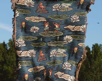 May include: A woven throw blanket featuring a dark blue background with a repeating pattern of orange frogs and white water lilies on green lily pads. The blanket has a fringed edge and a textured appearance with detailed illustrations of pond life.