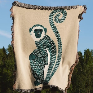 Może przedstawiać: Beżowy koc z motywem siedzącej małpy w kolorze morskim. Małpa ma białą twarz i morski kontur. Koc ma frędzel na krawędzi. Tło przedstawia błękitne niebo i zielone drzewa. Idealny do salonu.