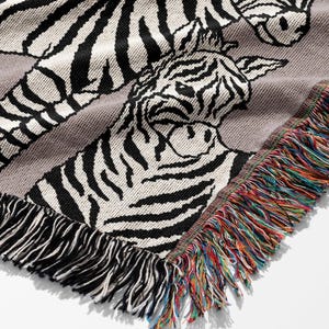 May include: A gray woven throw blanket featuring a zebra design. The blanket has black and white zebra stripes and a fringe border with a rainbow of colors. The blanket is made of a soft, woven material.