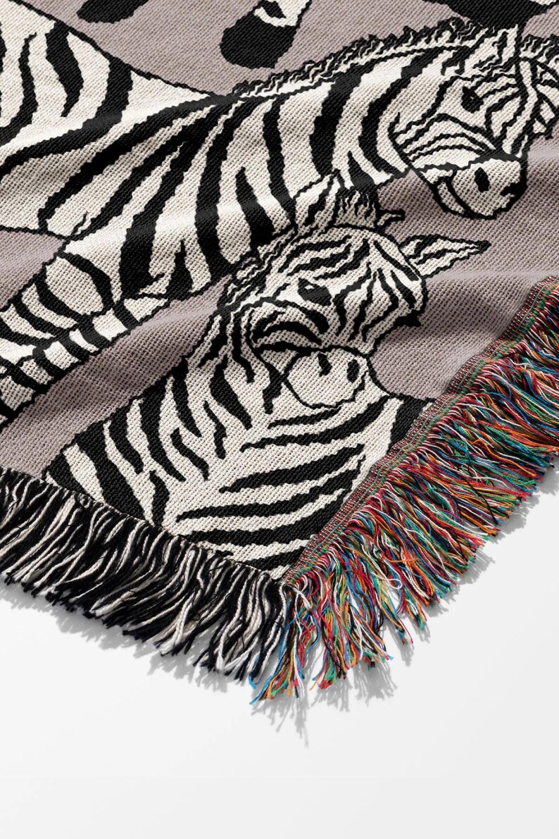 May include: A gray woven throw blanket featuring a zebra design. The blanket has black and white zebra stripes and a fringe border with a rainbow of colors. The blanket is made of a soft, woven material.