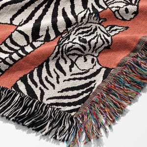 May include: A woven throw blanket featuring a repeating zebra pattern in black and white on a coral background. The blanket has a fringed edge with a rainbow of colors. The design is a close-up of the zebra's face.