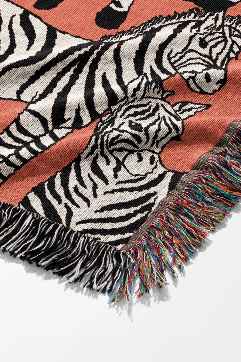 May include: A woven throw blanket featuring a repeating zebra pattern in black and white on a coral background. The blanket has a fringed edge with a rainbow of colors. The design is a close-up of the zebra's face.