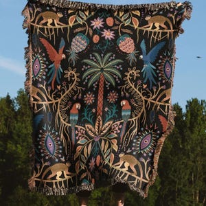 May include: A black woven throw blanket with a tropical design featuring parrots, monkeys, and palm trees. The blanket has a fringed edge and a detailed pattern in shades of brown, green, blue, and red. The blanket is held up against a blue sky.