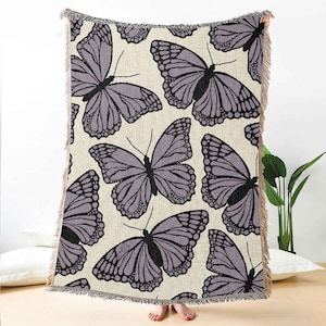 Puede incluir: Una manta tejida con un patrón repetido de grandes mariposas estilizadas en tonos grises y negros sobre un fondo crema. La manta tiene un borde con flecos y la sostiene una persona.