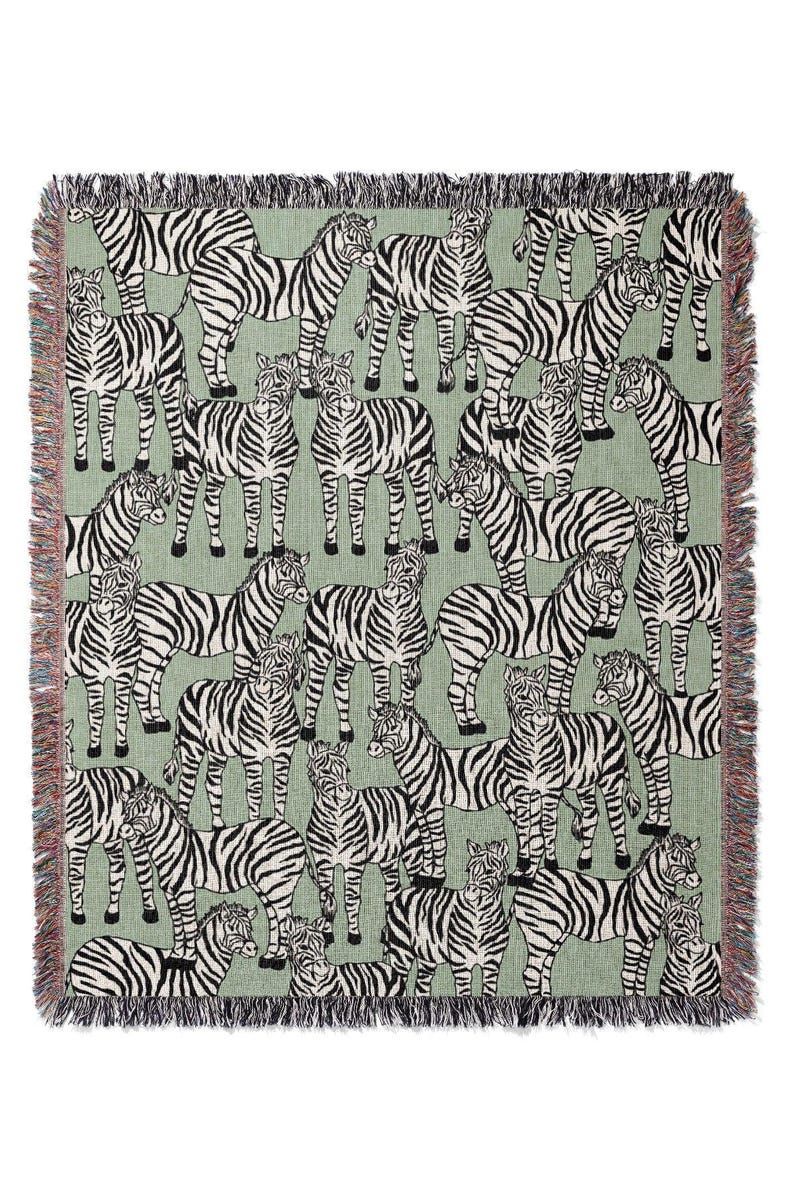 May include: A woven throw blanket featuring a repeating pattern of zebras in black and white on a light green background. The blanket has a fringed edge in various colors. This decorative blanket is suitable for a bed or sofa.