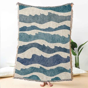May include: A woven throw blanket featuring a wave pattern in shades of blue and off-white. The blanket has a fringed edge and is held up, showcasing the design. The overall theme is coastal or nautical.