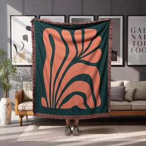 Puede incluir: Una manta grande y rectangular con un fondo verde oscuro y un diseño floral abstracto de color coral. La manta tiene un borde con flecos y se exhibe en interiores.