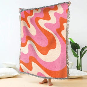 May include: A woven throw blanket featuring a retro wavy pattern in pink, orange, and cream. The blanket has a fringed edge. The design is reminiscent of the 1970s. The blanket is held up against a white background.