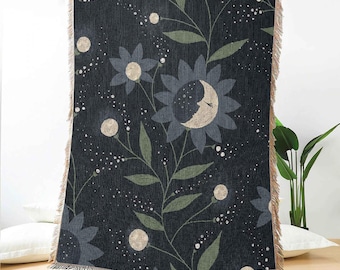 May include: A woven throw blanket featuring a dark charcoal background with a pattern of stylized flowers. The central flower has a crescent moon face with closed eyes. The design includes green leaves, small white dots, and fringed edges.