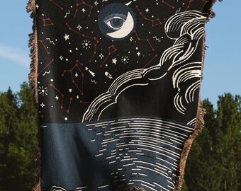 Può includere: Una coperta nera tessuta con un design celeste. La coperta presenta un cielo notturno con costellazioni, stelle e una luna crescente con un occhio. La metà inferiore ha un design ad acqua blu e bianco. I bordi hanno una frangia marrone.
