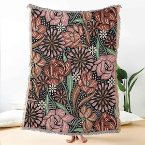May include: A woven throw blanket with a floral pattern. The design features large flowers in shades of brown, pink, and green against a black background. The blanket has fringed edges.