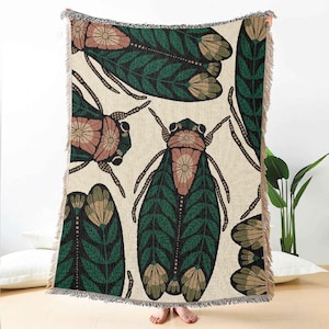 May include: Beige throw blanket with a repeating pattern of stylized insects. The insects feature green leaf-like wings, pink and beige floral accents, and brown antennae. The blanket has a fringed edge and is made of a woven fabric.
