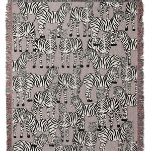 May include: A woven throw blanket featuring a repeating pattern of zebras in black and white on a gray background. The blanket has a fringed edge with a mix of colors. The design is a playful take on animal prints.