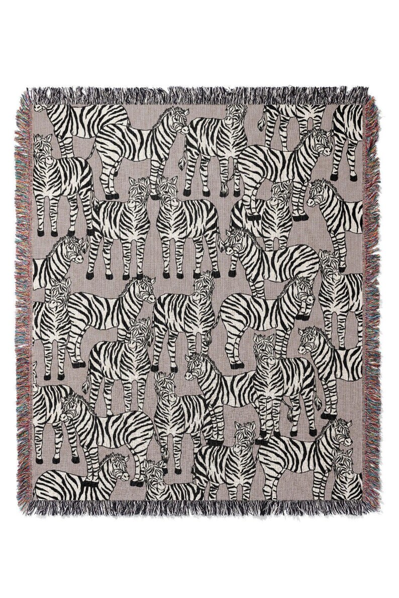 May include: A woven throw blanket featuring a repeating pattern of zebras in black and white on a gray background. The blanket has a fringed edge with a mix of colors. The design is a playful take on animal prints.