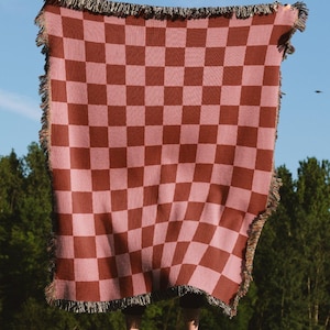 Può includere: Una coperta a quadri rosa e marrone con bordi sfrangiati è mostrata contro un cielo blu. Il motivo della coperta è un classico disegno a scacchiera. Questa coperta, adatta per picnic o decorazioni per la casa, è tenuta davanti a uno sfondo verde.