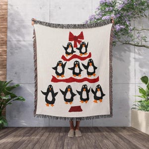 May include: Cream-coloured woven throw blanket featuring a Christmas tree design made of black and white penguins with orange feet. The tree has a red ribbon and base. The blanket has a fringed edge.