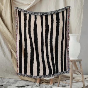 May include: A black and cream striped throw blanket with a fringed edge. The blanket features vertical stripes and is displayed against a neutral backdrop. The fringing has a mix of colours.