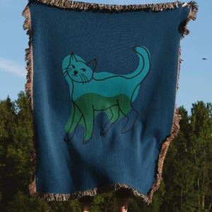 K&ouml;nnte beinhalten: Eine marineblaue Decke mit einer teal- und gr&uuml;nen Katzenillustration. Die Decke hat einen Fransenrand und wird vor blauem Himmel und gr&uuml;nen B&auml;umen gehalten.