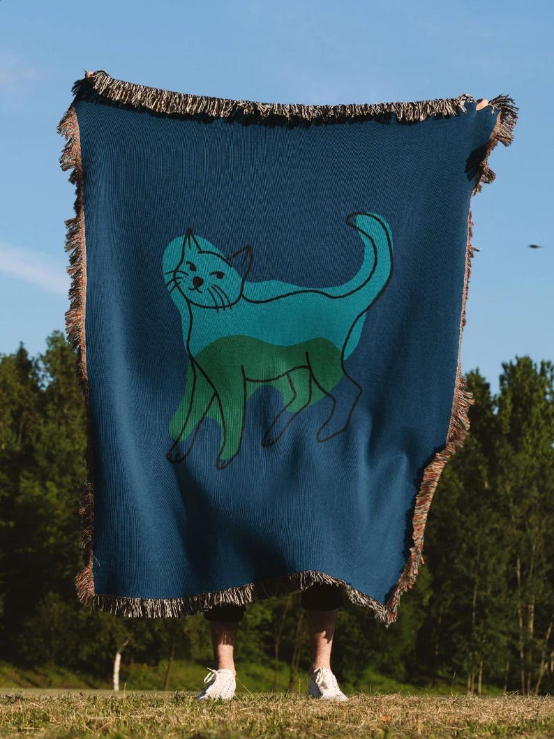 K&ouml;nnte beinhalten: Eine marineblaue Decke mit einer teal- und gr&uuml;nen Katzenillustration. Die Decke hat einen Fransenrand und wird vor blauem Himmel und gr&uuml;nen B&auml;umen gehalten.